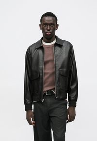 Casaco bomber de pele preta com dois bolsos frontais. Usado por cima de uma camisola texturada cor malva e calças cinzentas. Gola simples e fecho de correr.