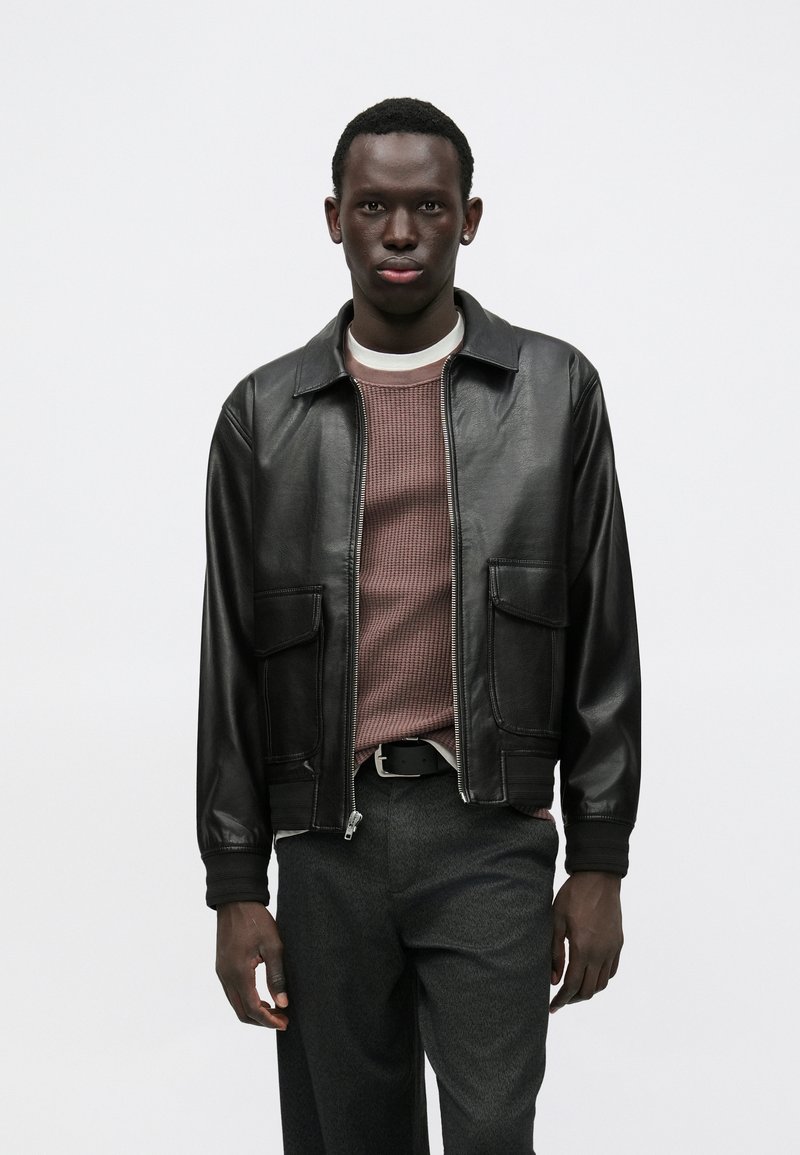 Casaco bomber de pele preta com dois bolsos frontais. Usado por cima de uma camisola texturada cor malva e calças cinzentas. Gola simples e fecho de correr.