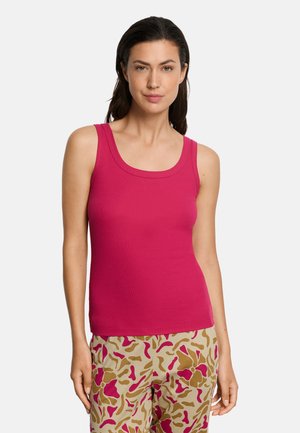 Vrouw draagt een mouwloze magenta tanktop en beige broek met roze en bruine abstracte patronen, staand tegen een witte achtergrond.