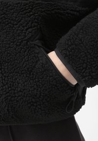 Pullover in pile nero con superficie testurizzata, dotato di una tasca con zip nella parte anteriore e di un orlo liscio a contrasto intorno al polso.