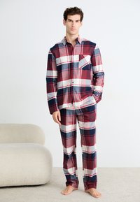 Hombre de pie descalzo, vistiendo una camisa de cuadros de manga larga en rojo, azul marino y blanco, y pantalones a juego, al lado de un sofá beige sobre una alfombra.