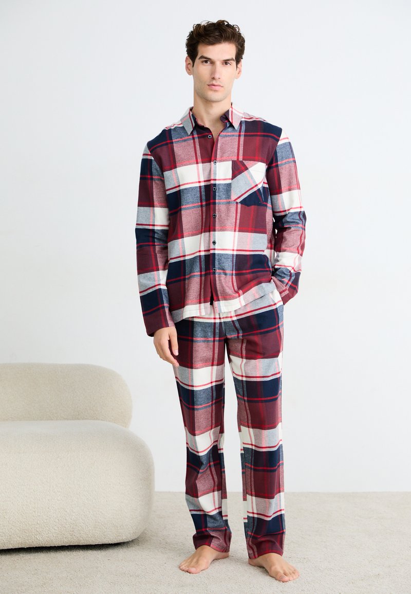 Hombre de pie descalzo, vistiendo una camisa de cuadros de manga larga en rojo, azul marino y blanco, y pantalones a juego, al lado de un sofá beige sobre una alfombra.