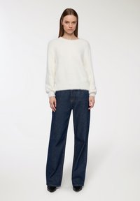 Biały puszysty sweter z długimi rękawami i prążkowanym dołem, w połączeniu z wysokimi ciemnymi dżinsami oraz czarnymi spiczastymi butami.