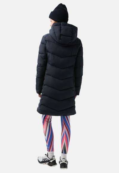 Dunkelblaues Puffer-Gemälde mit Wabenmuster und Kapuze, kombiniert mit mehrfarbigen gestreiften Leggings und schwarzen Turnschuhen.