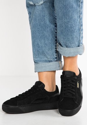 Person trägt hochgekrempelte blaue Jeans und schwarze Puma-Sneakers und steht auf einem hellen Boden vor einem schlichten Hintergrund.