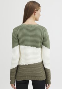 Pull à rayures vertes et blanches avec des bandes de textures variées, présentant un col rond et des poignets côtelés. Vu de dos, un jeans noir est visible.