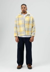 Gebreid jack met geruit patroon in geel en grijs, voorzien van een knoopsluiting, brede kraag en zachte textuur, gedragen over een wit T-shirt.