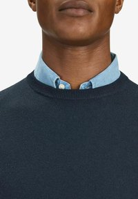 Marinblå crew-neck tröja med en slät yta, lagd över en ljusblå skjorta med krage. Har ribbade ärmslut och nederkant.