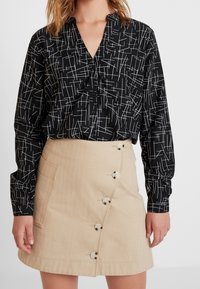 Camicia nera con bottoni e linee astratte bianche, abbinata a una gonna beige fino al ginocchio con chiusura a bottoni laterale e tasca.