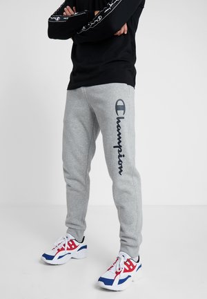 Osoba v šedých teplácích značky Champion a bílých teniskách s modrými a červenými akcenty stojící se založenýma rukama.