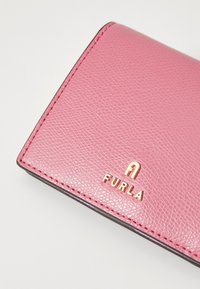 Ružičasti kožni novčanik s teksturiranom površinom, s zlatnim logotipom "FURLA". Rubovi su tamne boje, ističući dizajn.