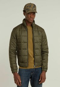 Olivgrön puffjacka med quiltat mönster, dragkedja och ribbade manchetter, ihop med en brun skjorta och denimjeans. Kanske har kepsen ett kamouflagemönster.