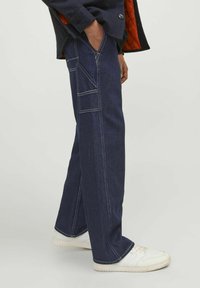 Temno modri denim hlače s ravnimi nogavicami, ki imajo bele šive in stranske žepe, v kombinaciji z belimi supergami.