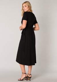 Zwarte jurk tot de knie met korte mouwen, een strikdetail in de taille en een soepele, vloeiende textuur. Gecombineerd met zwarte strap-sandalen.