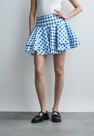 Γυναίκα που φοράει μπλε και λευκή καρό φούστα με ανοιχτή όψη, λευκό αμάνικο μπλουζάκι και μαύρα ίσια παπούτσια, στέκεται μπροστά σε απλό φωτεινό φόντο.