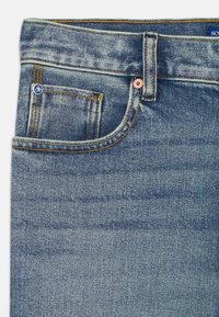Jeans in denim di colore blu medio con cuciture arancioni a contrasto, un bottone di rame e un piccolo etichetta con il logo sulla tasca anteriore.