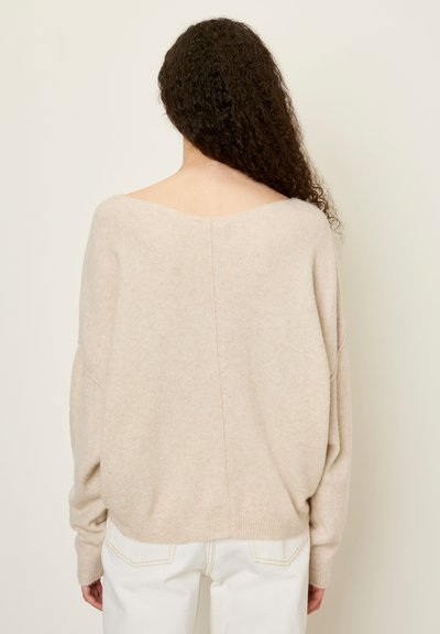 Femme aux longs cheveux bouclés portant un pull ample beige avec un large décolleté dans le dos et un pantalon blanc, debout devant un fond uni.