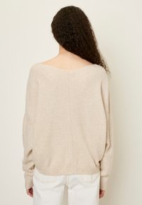 Beige oversized trui met een brede V-hals en een drop shoulder ontwerp. Zachte textuur, geribbelde manchetten en subtiele naaddetails aan de achterkant.