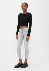 Γυναίκα με μαύρο μακρυμάνικο κοντό τοπ, ανοιχτό γκρι στενά τζιν και μαύρα μοκασίνια, στέκεται μπροστά σε απλό λευκό φόντο.