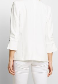 Blazer bianco leggero con maniche a 3/4 svasate, dettagli arricciati sottili sulle maniche e un design posteriore pulito e su misura.