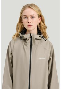 Ung kvinde med bølget blondt hår iført en beige Tretorn regnjakke med lynlås foran og justerbar hætte.