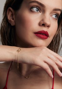 Złote kolczyki i bransoletka w kształcie serca, z delikatnymi wzorami łańcuszka. Bogate czerwone usta i czerwona bluzka uzupełniają biżuterię.