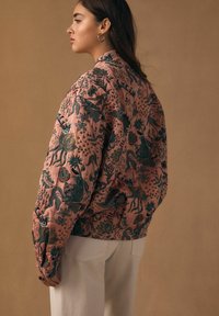 Gewatteerd bomberjack in lichtroze met ingewikkelde donkere bloemen- en dierenpatronen. Heeft een ontspannen pasvorm en geribbelde boorden.