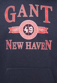 Sudadera de color marinero que presenta un texto rojo intenso: "GANT", "NEW HAVEN" y un emblema circular con "49" y "1949". Tejido suave, bolsillo tipo canguro.