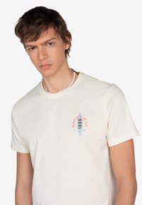 Camiseta de algodón color crema con un gráfico de tabla de surf azul y texto rojo. Las mangas cortas y el escote de crew completan el diseño.