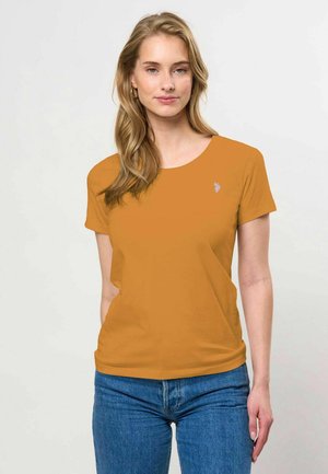 Femme aux longs cheveux blonds portant des boucles d'oreilles créoles dorées, un t-shirt jaune moutarde à manches courtes et un jean bleu sur un fond clair uni.