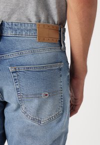 Pohled zezadu na osobu v světle modrých džínech Tommy Jeans s béžovou koženou značkovou nášivkou a malou vlajkovou logem na kapse, šedé tričko zasunuté dovnitř.