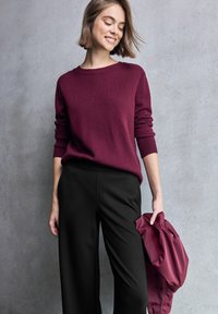 Bordeaux trui met geribbelde manchetten en een ontspannen fit, gecombineerd met zwarte tailor-made broek, terwijl hij een bijpassende bordeaux jas vasthoudt. Gladde texturen.