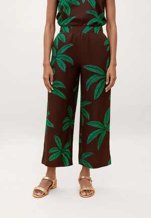 Pantalones de pierna ancha marrones con estampado de grandes hojas tropicales verdes, combinados con sandalias planas doradas sobre fondo claro.