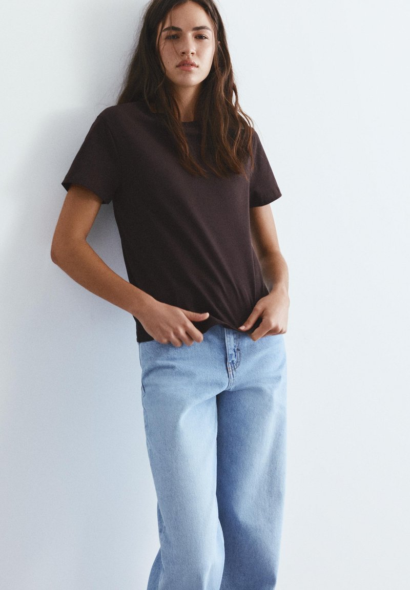 Jeune femme aux longs cheveux foncés portant un t-shirt noir et un jean bleu clair, debout contre un mur blanc uni.