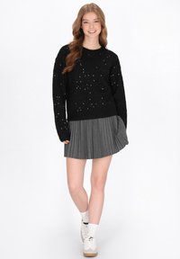 Fekete, texturált pulóver ezüst díszítéssel, amelyet egy pliszírozott szürke szoknyával és fehér cipővel párosítottak. Az outfit laza szabású és kerek nyakú.