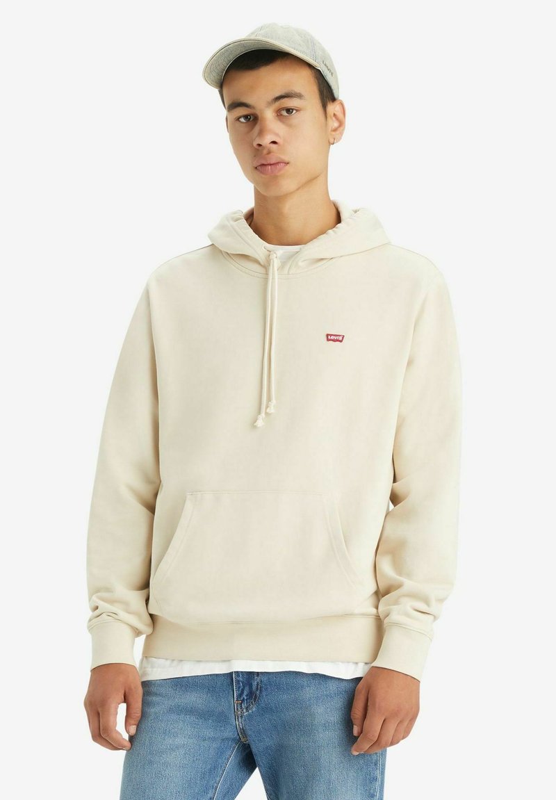 Cream-colored hoodie made of cotton, features a front pocket, drawstring hood, and a small red logo. Worn with light blue jeans.