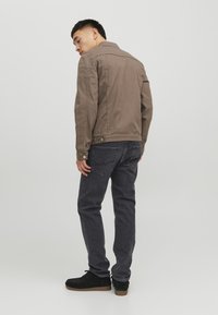 Giacca in tela marrone con colletto, tasche laterali e dettagli con bottoni, abbinata a jeans grigio scuro e scarpe in suede nere.