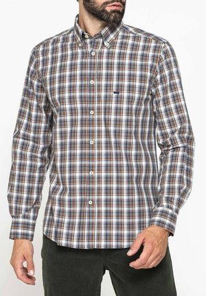 Uomo che indossa una camicia a quadri a maniche lunghe abbottonata con tonalità marroni, grigie e bianche, abbinata a pantaloni verde scuro.