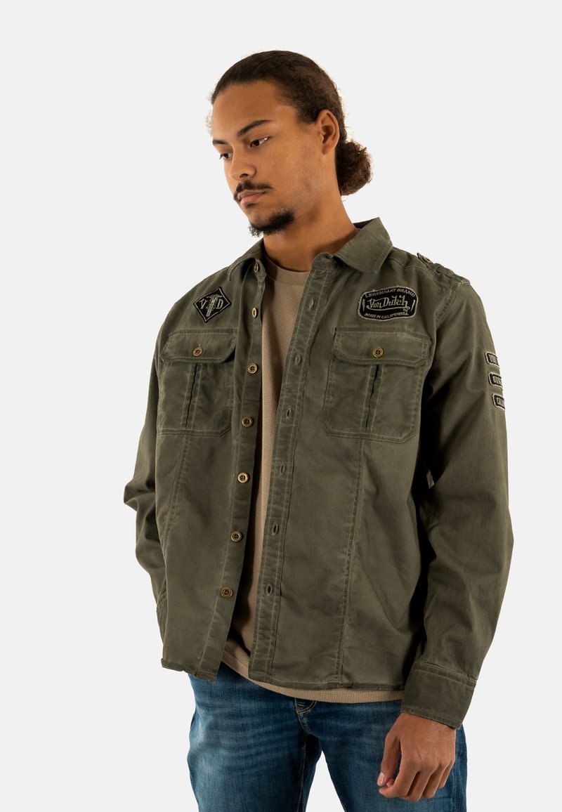 Giacca camicia in tela verde oliva con due tasche sul petto, chiusura a bottoni e toppe ricamate. Abbinata a una maglietta beige e jeans blu.