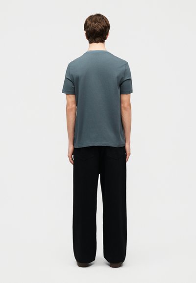 Άνδρας με κοντά καστανά μαλλιά, που φοράει μπλε πουκάμισο με κοντά μανίκια και μαύρο παντελόνι με φαρδιά πόδια, στέκεται με την πλάτη προς τα εμπρός σε ένα απλό φόντο.