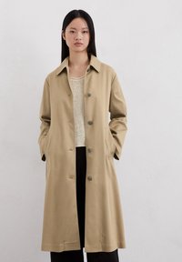 Cappotto lungo beige in tessuto morbido con colletto, chiusura frontale con bottoni, tasche laterali e vestibilità comoda.