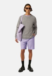 Grå struktureret sweater, lavendel shorts, sorte kraftige sko og matchende tote bag; model bærer mørke solbriller og sorte sokker.