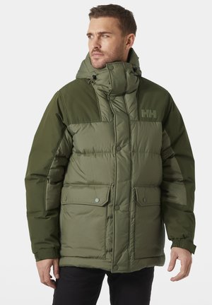 Hombre con chaqueta de invierno acolchada de color verde oliva, con cuello alto, capucha, botones de presión y dos bolsillos frontales con solapa, mirando hacia la izquierda.