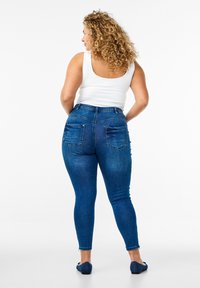 Jean en denim bleu taille haute avec un design ajusté, deux poches arrière et une texture lisse, associé à un débardeur blanc.