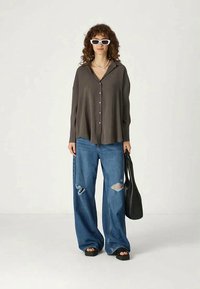 Camisa cinza com botões, jeans azuis rasgados e largos, sandálias pretas e uma mala tote preta. Óculos de sol retangulares brancos e cabelo encaracolado.