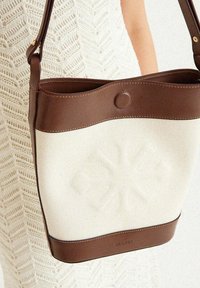 Braune Ledertasche mit einem cremefarbenen Canvas-Körper, der ein strukturiertes Design und eine abgerundete Oberseite aufweist. Mit passendem Lederbesatz akzentuiert.