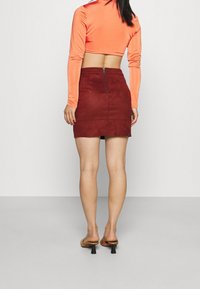 Mini-jupe en faux suède bordeaux avec une fermeture éclair à l'arrière. Associée à un crop top à manches longues orange et des sandales à talons brunes.