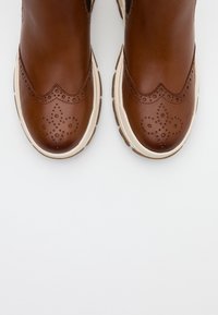 Botas de cuero marrón hasta el tobillo con detalles en relieve y un diseño en forma de olas. La suela es de color crema con acentos texturizados.