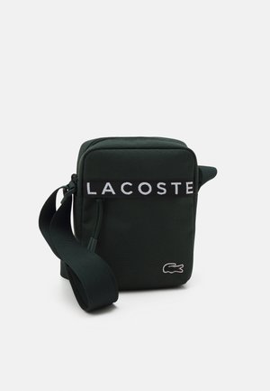 Sac bandoulière vert foncé avec une fermeture éclair, comprenant une bande noire marquée "LACOSTE" et un petit logo brodé en rouge et blanc.