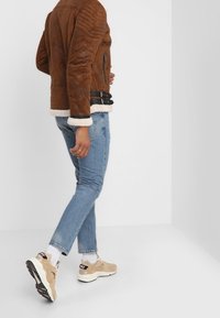 Brun læderjakke med quiltede detaljer, hvid shearlingforing, sorte accenter, lyseblå jeans og brune sneakers med sort logo.